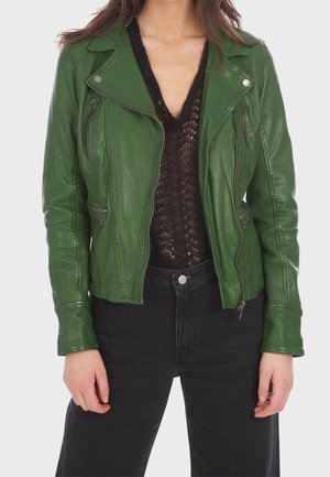 Femme portant une veste en cuir verte sur un haut en dentelle noire et un pantalon noir taille haute, debout les mains détendues le long du corps.