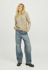 Felpa beige a maglia con cappuccio e cordino, indossata con jeans ampi blu chiaro. Stivaletti alla caviglia marrone scuro completano l'outfit casual.