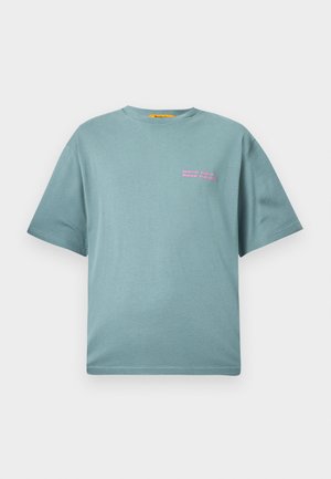 Vaaleansininen puuvillapaita, jossa on rento istuvuus. Ominaisuuksiin kuuluu pyöreä kaulus ja lyhyet hihat. Vasemman rinnan kohdalla on vaaleanpunaisella tekstillä "Tee kovasti töitä, juhli kovasti".