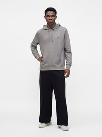 Jeune homme debout portant un sweat à capuche gris clair, un pantalon noir large et des baskets blanches sur fond blanc uni.