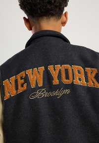 Person wearing a black jacket with large orange "NEW YORK" and smaller yellow "Brooklyn" embroidered on the back, facing away.