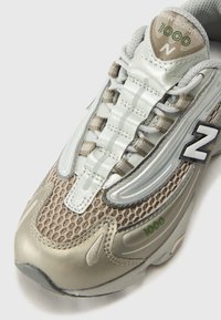Baskets New Balance avec une tige en mesh et synthétique, couleurs gris et beige, accents réfléchissants, et un logo "1000" bien visible sur le côté.