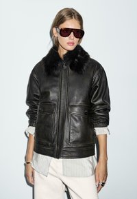 Schwarze Lederjacke mit einem Kragen aus Kunstfell, zwei vorderen Taschen und elastischen Bündchen. Kombiniert mit übergroßen, dunklen Sonnenbrillen und einem hellgrauen Shirt.