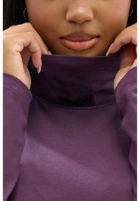 Pull à col roulé violet avec une texture douce, tissu légèrement froncé au niveau du cou et coupe décontractée, partiellement couvert par les mains.