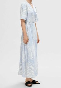 Dlouhé pruhované šaty s výstřihem do V, krátkými rukávy a páskem v pase. Modro-bílý vzor, lehká tkanina, nošeny s černými sandály.