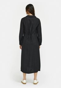 Svart långärmad klänning med figurnära midja och rak silhuett, tillverkad av lätt tyg, med en klassisk krage och elastiska rynkor.