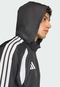 Μαύρο μπουφάν με κουκούλα κατασκευασμένο από ελαφρύ υλικό, με λευκές ρίγες κατά μήκος των μανικιών και λογότυπο στο στήθος.