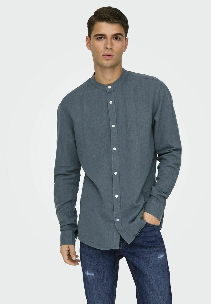Camisa gris de botón con cuello mao, mangas largas y textura tejida, combinada con jeans oscuros desgastados.