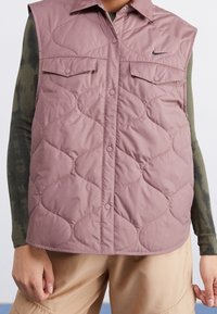 Světle růžová prošívaná vesta s límečkem, patentkami, dvěma předními kapsami a černým logem. Nosená přes tmavě zelenou košili s dlouhým rukávem.
