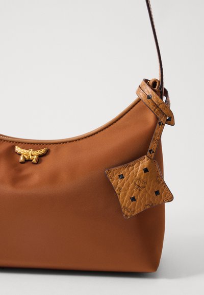 Brun läderhandväska med en böjd topp, guldvingad logotyp och mönstrad läderetikett hängande från remmen mot en vit bakgrund.