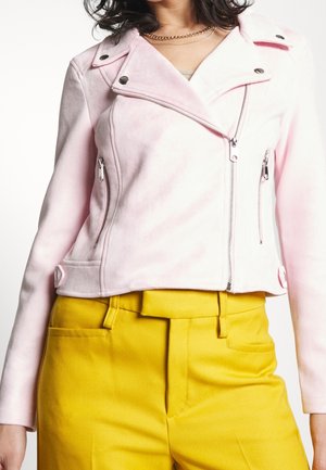 Femme portant une veste biker rose clair zippée associée à un pantalon taille haute jaune vif, montrée du cou aux hanches.