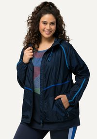 Marinblå vindjacka med blå accenter, lättviktig, har huva, blixtlåsfickor och en strukturerad yta. Lager på en mönstrad topp.