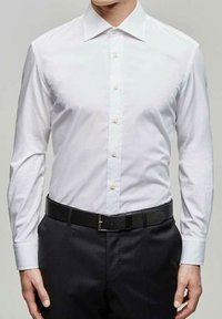 Chemise blanche à manches longues avec un col ouvert, associée à un pantalon sombre et une ceinture noire avec une boucle en argent.
