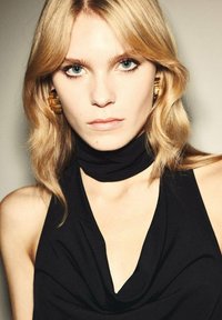 Čierny top bez rukávov s ozdobným výstrihom a vysokým golierom. Modelka má svetlo blond vlasy a nosí veľké textúrované zlaté náušnice.