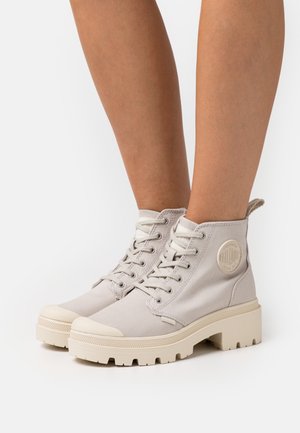Beige canvas ankelstövlar med texturerad yta, rund tå, snörning och gummisula. Har en rund logotypdetalj på sidan.