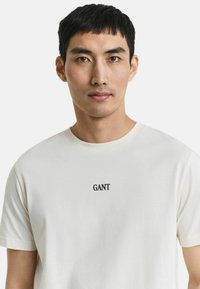 Uomo con capelli neri corti che indossa una maglietta bianca GANT su uno sfondo chiaro e uniforme, rivolto in avanti con un'espressione neutra.