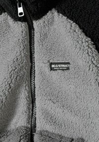 Veste zippée en polaire noire et grise. Elle présente un tissu texturé et doux avec une fermeture éclair centrale et une étiquette cousue portant la mention "DE-C/STRUCT."