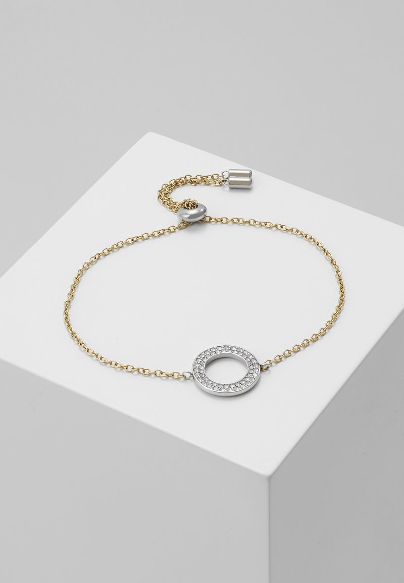 Bracelet délicat en chaîne d'or avec un pendentif circulaire en argent incrusté de petites pierres claires reposant sur un bloc d'exposition blanc.