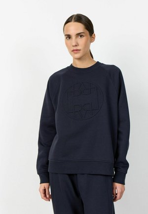 Kobieta z ciemnymi włosami związanymi do tyłu, ubrana w granatową bluzę i pasujące spodnie na jednolitym białym tle.