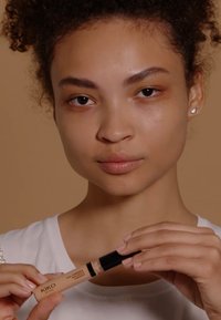 KIKO concealer i en plastiktube med sort låg. Modellen holder den og har en hvid skjorte på. Lysebrun baggrund, glat hudtekstur.