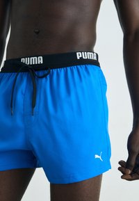 Mėlyni maudymosi šortai iš lengvos medžiagos, su juodu liemeniu, ant kurio yra baltas "PUMA" logotipas, juoda raištelio virvelė ir mažas baltas logotipo akcentas.