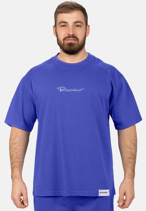 Mann mit kurzen Haaren und Bart, der ein lockeres blaues T-Shirt mit weißem Text trägt und vor einem schlichten hellen Hintergrund steht.