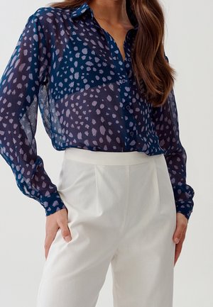 Kvinde iført en gennemsigtig navyblå bluse med lyse lilla abstrakte pletter, kombineret med højtaljede hvide skræddersyede bukser, stående med hænderne i lommerne.