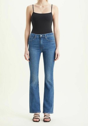 Kvinde iført en sort spaghetti-strop top, blå højttaljede jeans med let vidde og sorte sandaler med remme, stående mod en ensfarvet baggrund.