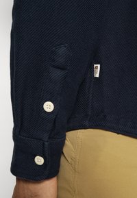 Marinblå skjorta med texturerat tyg, ärmar med manschetter som har krämfärgade knappar, samt en liten märkesetikett på sidan.