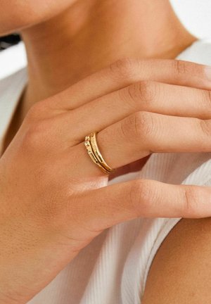 Zelta krāsas gredzenu komplekts ar trim joslām, kurām ir teksturēts, viļņots dizains un mazi caurspīdīgi akmentiņi uz vienas joslas.