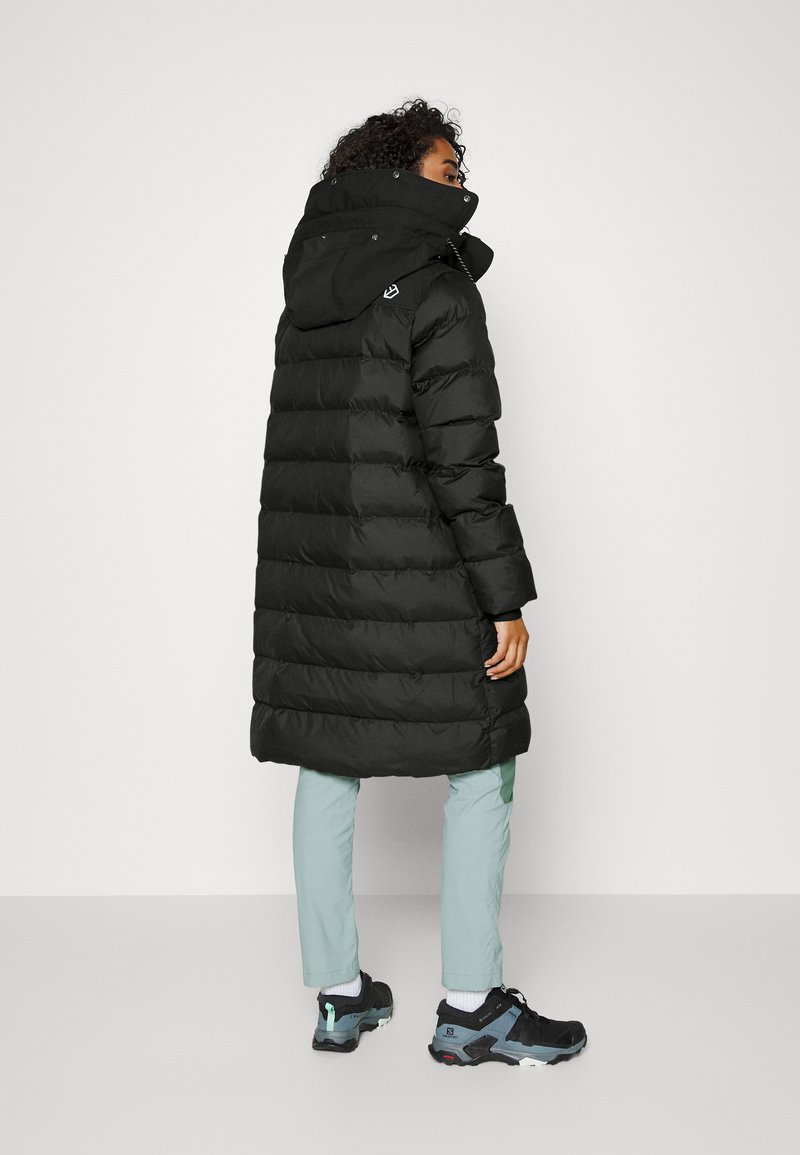 Manteau en duvet noir avec capuche, doté d'un design matelassé et d'une forme légèrement allongée. Porté avec un pantalon vert clair et des baskets noires.