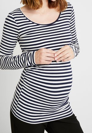 Gravid kvinde iført en sort og hvid stribet langærmet skjorte, hænderne hviler blidt på maven.