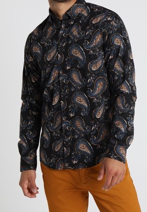 Homme portant une chemise noire à manches longues avec des motifs cachemire marron et bleu et un pantalon couleur moutarde.