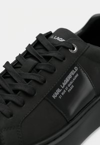 Melnas sporta kurpes ar gludu ādas virsu, plakaniem melniem šņorēm un spīdīgu zīmola etiķeti sānos. Noapaļots pirksts un minimālisma dizains.