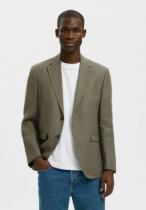 Młody mężczyzna stoi, ubrany w oliwkową marynarkę na białej koszulce i niebieskich dżinsach, z lewą ręką w kieszeni na jednolitym tle.