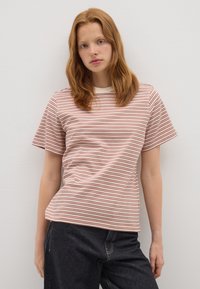 Mujer joven con cabello rojo hasta los hombros, vistiendo una camiseta a rayas blanca y marrón y pantalones oscuros, de pie contra una pared blanca lisa.