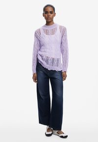 Lila gestrickter Pullover mit offenem Maschenmuster, langen Ärmeln und asymmetrischem Saum, kombiniert mit weit geschnittenen dunklen Jeans und flachem Schuhwerk.