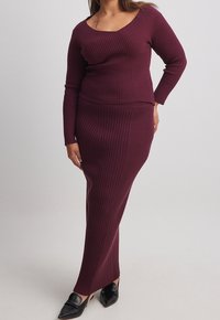 Burgundy ribbad stickad topp med långa ärmar och matchande lång kjol; figurnära silhuett; slät textur; spetsiga svarta skor.