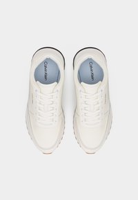 Ζευγάρι λευκά αθλητικά παπούτσια Calvin Klein με κορδόνια, ορατό λογότυπο στις πλευρές και στις εσωτερικές σόλες, εμφανιζόμενα από πάνω σε απλό λευκό φόντο.