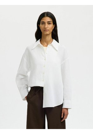 Femme aux cheveux longs et foncés portant une chemise blanche ample à boutons et un pantalon marron foncé, debout devant un fond uni.