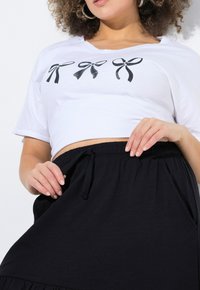 Weißes, kurz geschnittenes T-Shirt mit drei schwarzen Schleifen-Designs, kombiniert mit einem schwarzen Rock mit elastischem Bund und seitlichen Taschen.