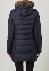 Giacca piumino blu navy con cappuccio e rifinitura in pelliccia, caratterizzata da cuciture verticali e un design aderente, che arriva fino a metà coscia. Tessuto testurizzato.