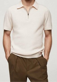 Homme portant un polo en tricot beige à manches courtes avec fermeture éclair, rentré dans un pantalon marron plié taille haute, les mains dans les poches, sur fond uni.