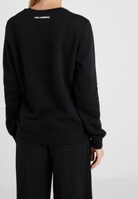 Svart sweatshirt med rundad halsringning, ribbade ärmslut och nederkant. Logotypdetalj på övre delen av ryggen. Materialet ser mjukt och texturerat ut.