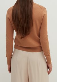 Jasnobrązowy, dopasowany sweter z długimi rękawami, prążkowanymi mankietami i miękką fakturą, zestawiony z jasnobeżowymi spodniami dla neutralnego wyglądu.