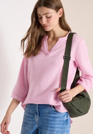 Femme portant un haut rose clair à manches longues et un jean bleu, tenant un sac à bandoulière matelassé vert foncé avec une large sangle.