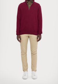 Bordeauxfarbener ZIP-Pullover über einem karierten grünen Hemd, kombiniert mit beigen Hosen und weißen Sneakers. Glatte Textur, lässiges Design.