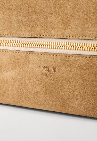 Smeđi velur torba s zlatnim detaljima na zatvaraču. Ima urezani logotip "Moschino Milano". Glatka tekstura s šavovima.