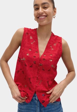 Mujer sonriente con blusa roja sin mangas de encaje abotonada y jeans azules, con las manos apoyadas cerca de los bolsillos sobre un fondo liso.