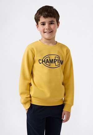Sudadera amarilla de tela suave con cuello redondo, que presenta un logo negro de "CHAMPION" en el pecho. Usada con pantalones oscuros.